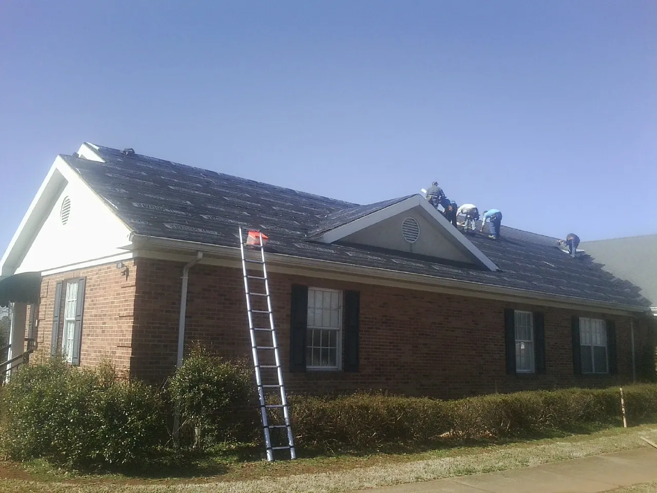 Crew tearing shingles off a brick ranch roof with a ladder set — Metal Roof Repair work by our crew in Bigelow