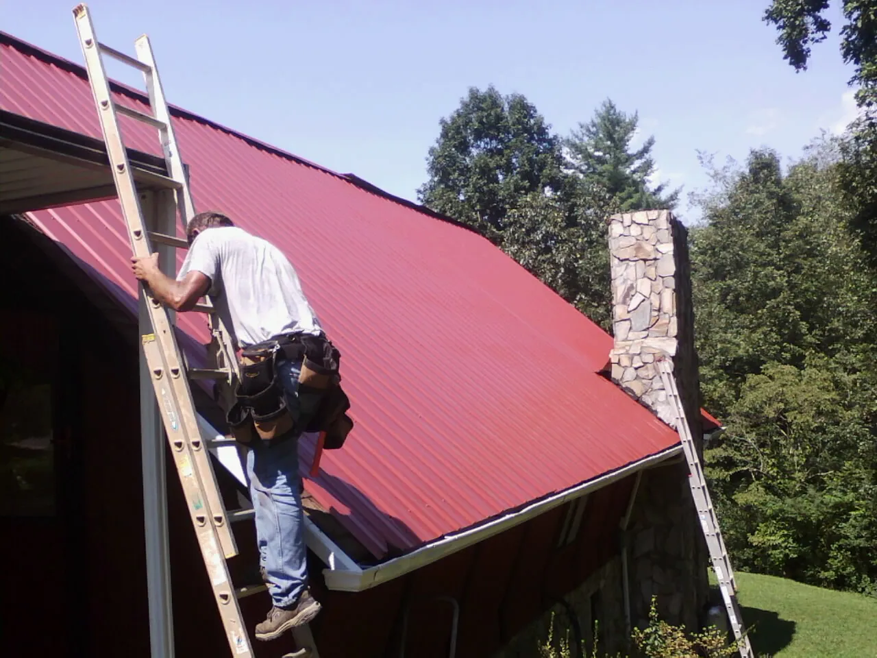 Roofer on a ladder beside a red metal roof and stone chimney — Soffit & Fascia Repair by Roofing Brothers in Bigelow
